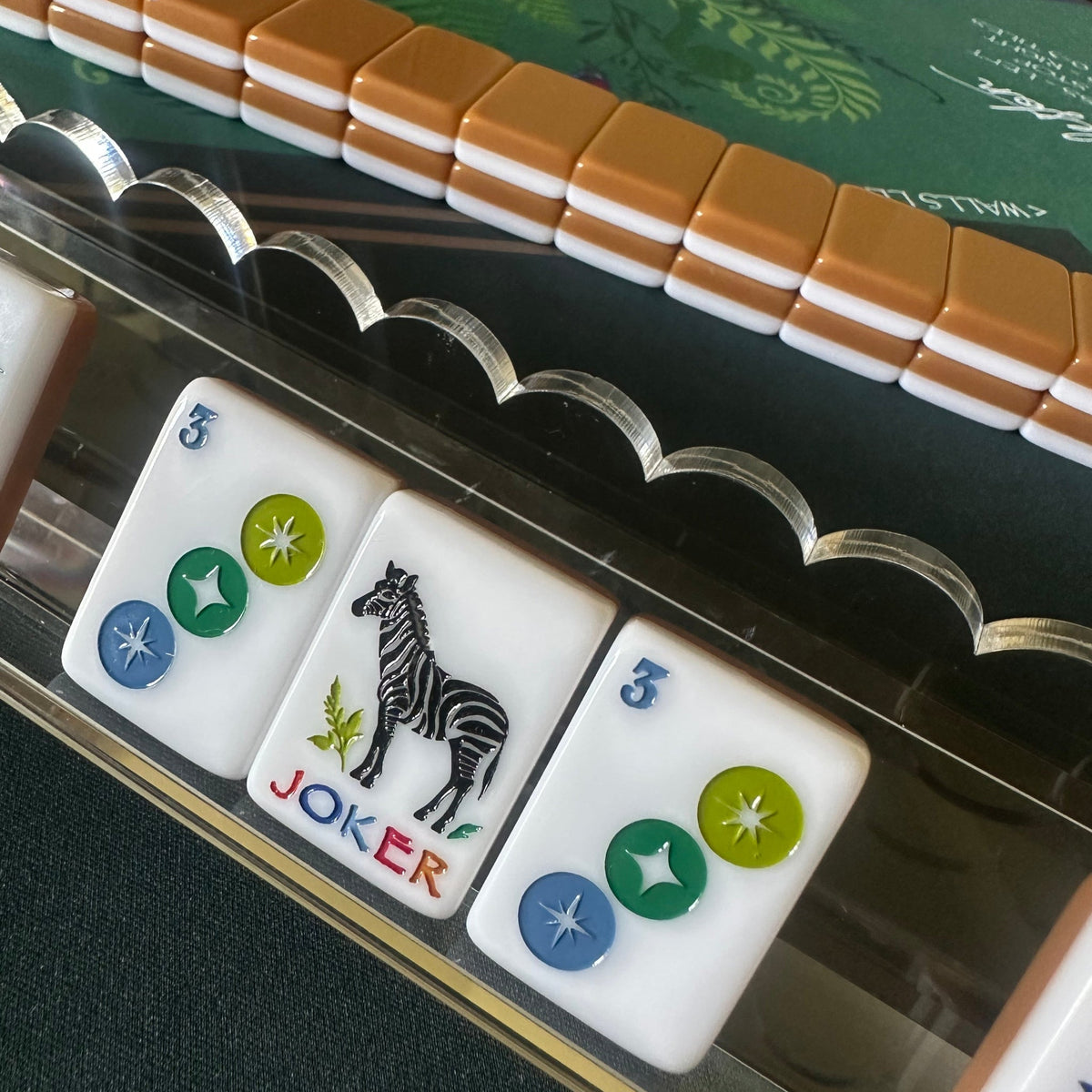 Clear acrylic mahjong rack with a scalloped-edge pusher holding colorful tiles on a dark green playing mat